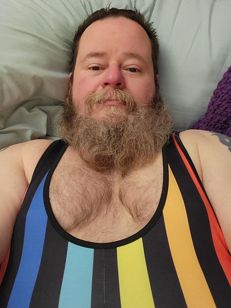 A bearded man with a hairy chest wearing a rainbow singlet looks up from his bed.