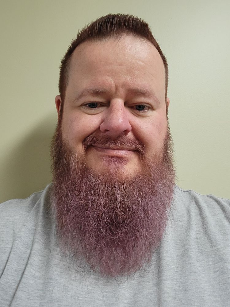 middle aged man with blue eyes, brown hair and beard dyed purple.  fresh short haircut and some shaping on the large beard.