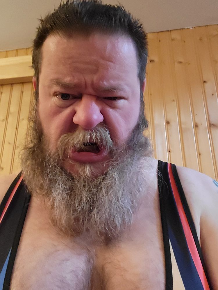 A bearded man with a hairy chest wearing a rainbow singlet makes a snarly face at the camera.