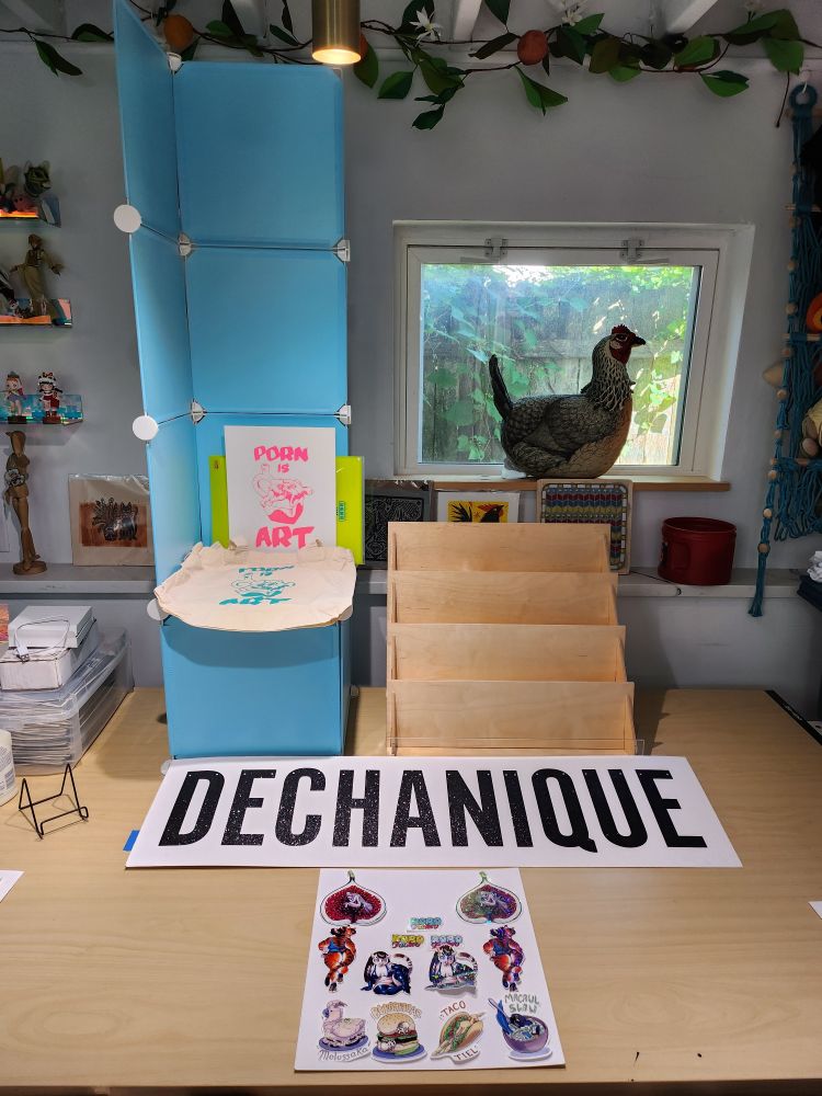SPX display in progress! A blue print tower and an empty book rack on a wood table. A big sign reads "Dechanique" while a smaller sign of stickers is being laid out. A tote bag and print reading "porn is art" sit on the display