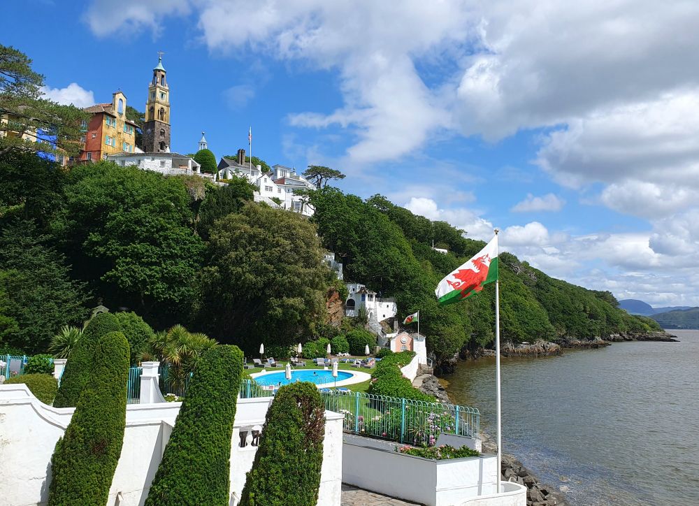 View from The Hotel showing the dramatic location of Portmeirion village perched high above the estuary of the River Dwyryd. The distinctive Campanile is a landmark on the skyline.
In the foreground are topiaried trees and a flagpole flying 'Y Ddraig Goch' (the red dragon), the flag of Wales.
The Blue oval of an outdoor swimming pool is visible at the base of the hillside.