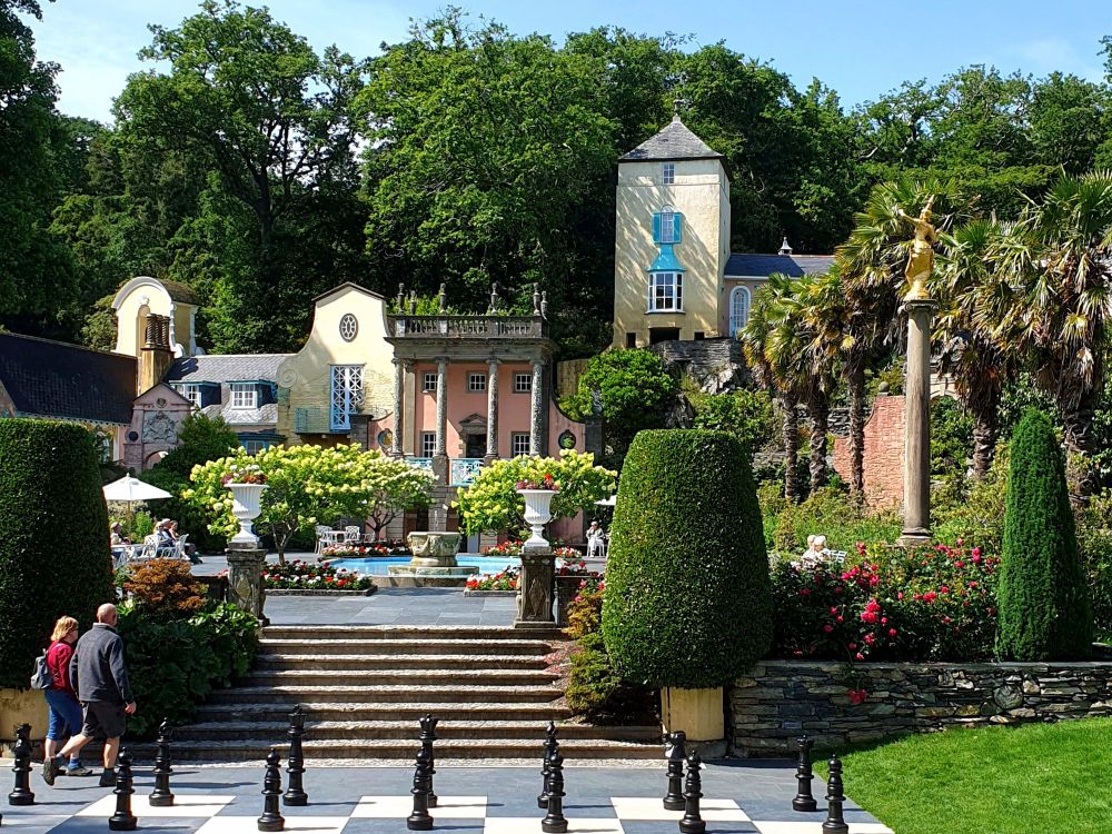View across to the Central Piazza at Portmeirion. In the foreground is a giant chessboard. Stone steps lead up to a a paved, and beyond that a fountain pool and the Gothic Pavilion. 