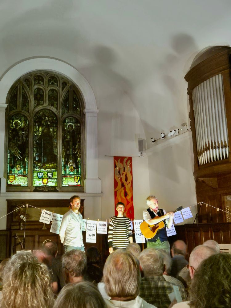 Three performers preparing for the closing song in the show 'Three Acres + a Cow'. They are standing in front of a washing line hung with paper banners printed with the dates and titles of each of the chapters in the history of land rights in Britain from 1066 to the present day.