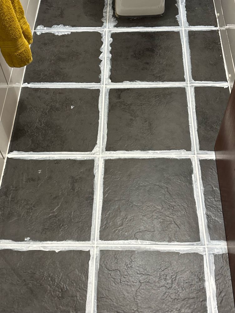 Bathroom floor with black tiles and white thick lines between them, caused by grout reviver. The tiles are not centred to the room, and instead are fitted 2.5 across, looking very wonky. 