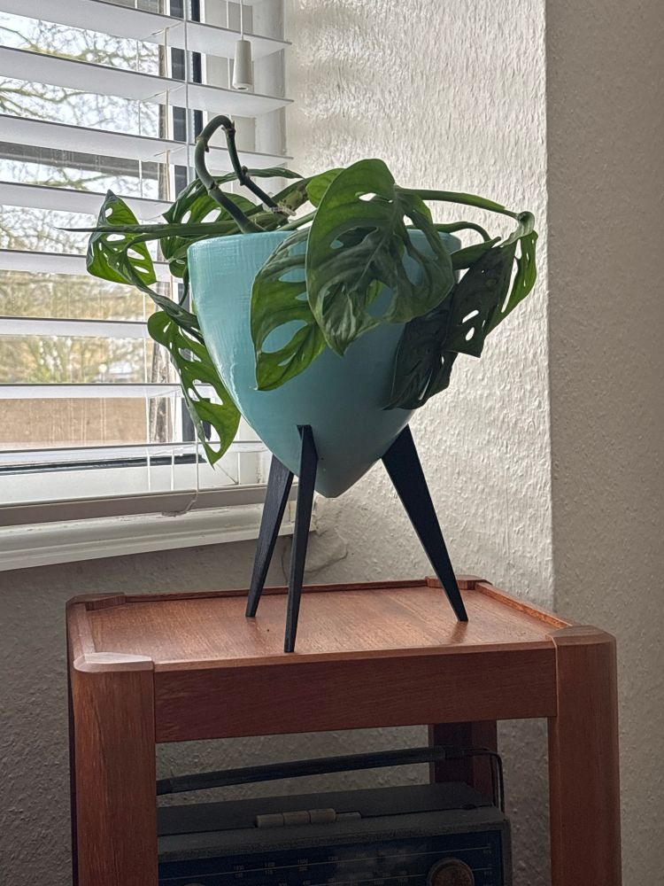 An aqua coloured cone-shaped plant pot on three black tripod legs sits on top of a slim, square, teak display cabinet in front of a window with a white Venetian blind. 