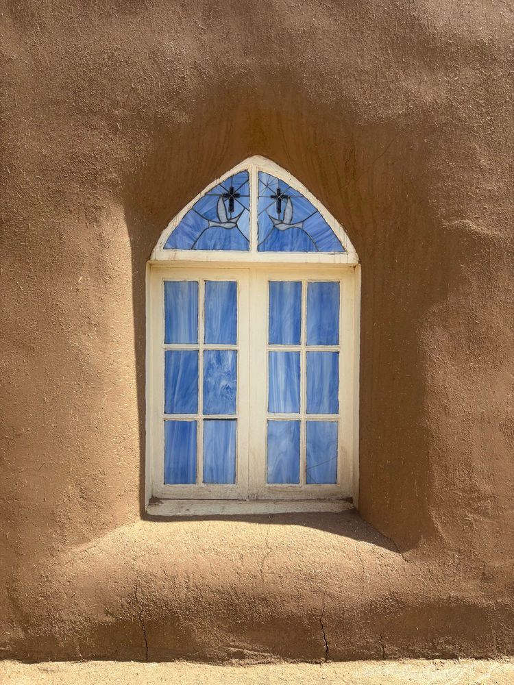Stained glass window of the church in Taos Pueblo. The white window frame is offset by sky blue panes, the top triangular panels each have a dove and cross