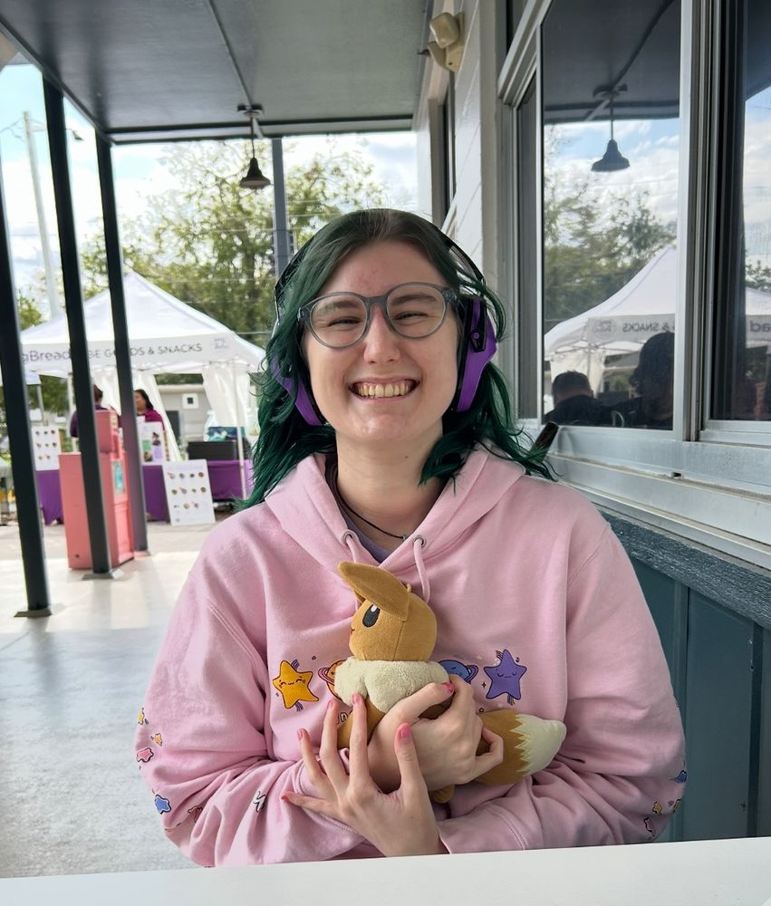 lohla, a white person with green hair, sitting outside smiling. they are wearing a pink sweatshirt, purple ear defenders, and are holding their eevee plush