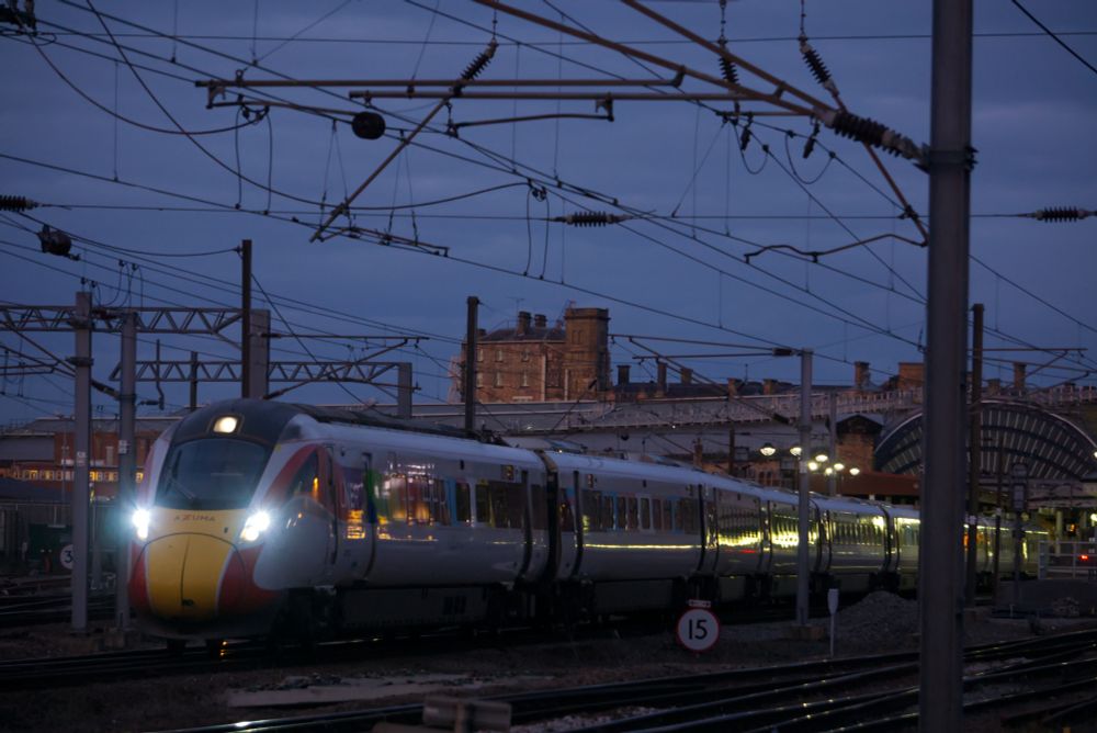 An electric high speed train departs a victorian built railway station with its iconic curved train shed. The train features a livery with both the standard LNER colour scheme (white with a red stripe along its length and yellow at the very front below the cab window), but with a different pride flag found on each coach within that red stripe.