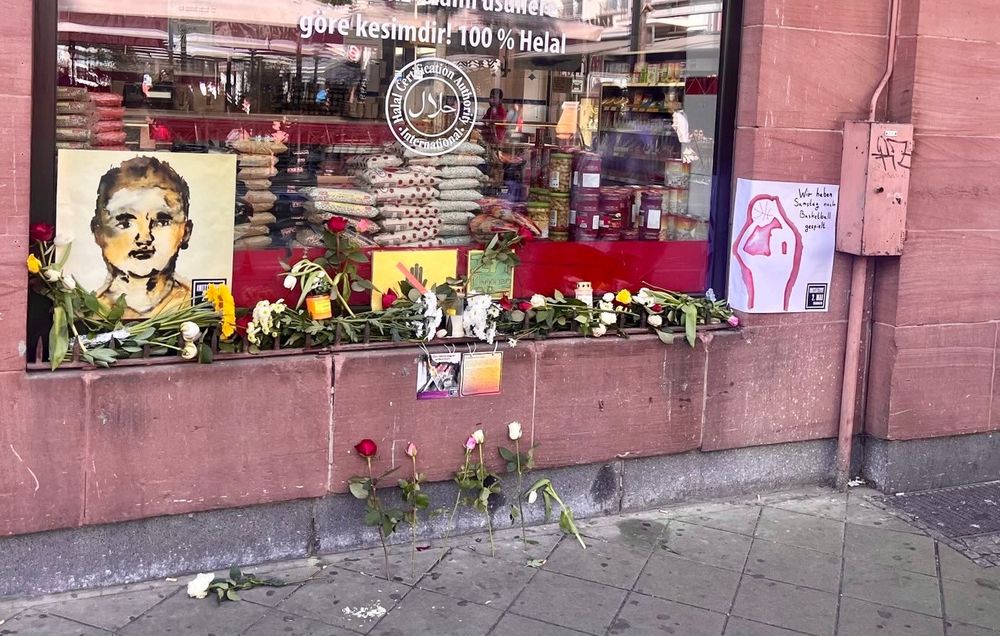 Vor dem Schaufenster der Metzgerei befindet sich ein Metallsteg mit nach oben ragenden Zacken. Zwischen den Metallstegen befinden sich unendlich viele verschiedene Schnittblumen und echte und auch batteriebetriebene Grablichter Auf dem Boden, an der Mauer angelehnt, nochmal 7 Rosen. Die fanden keinen Platz mehr zwischen den Metallstegen.
Auf dem Schaufenster rechts, mittig, links vom Schaufenster und an der Mauer unterhalb des Schaufensters 4 Auqarellbilder. Eines (DIN A 3) mit Antes Kopf, andere die Infos zu Ante enthalten und eines „In Memoriam“. Dazu noch auf gelbem Papier eine Hand, die mit dickem roten Strich durchgestrichen ist. 
