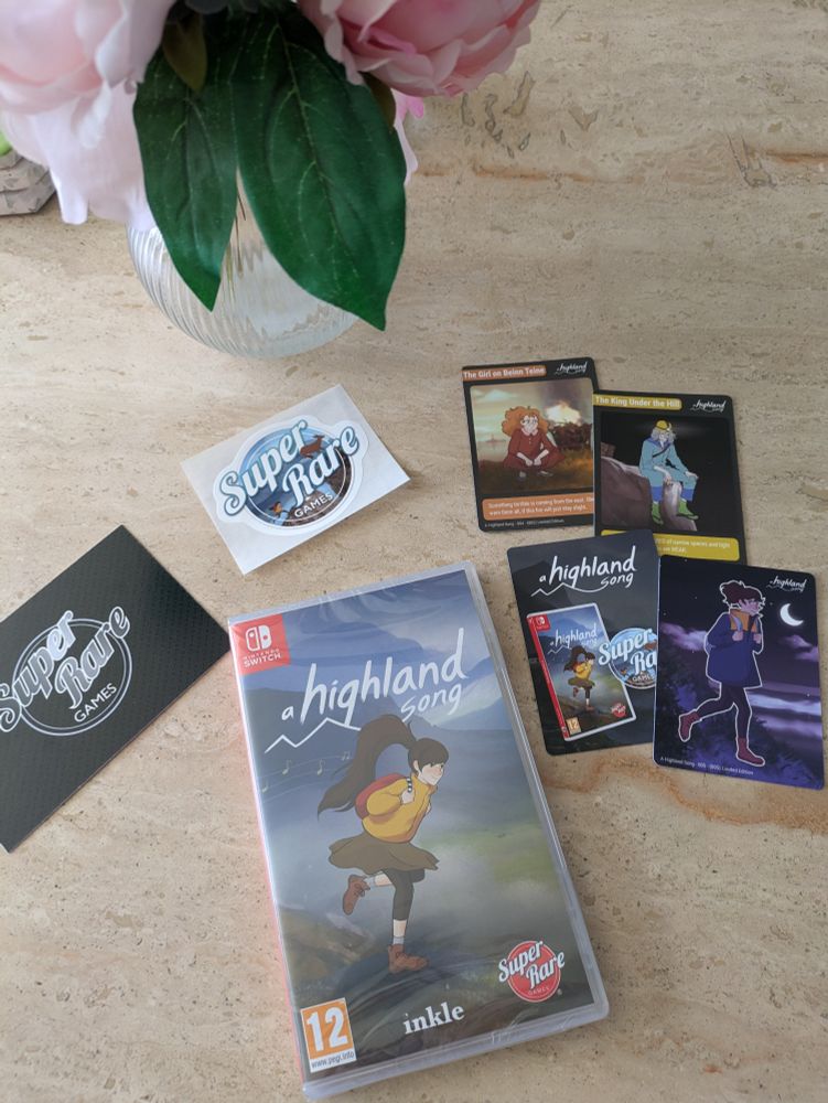 An unopened physical copy of A Highland Song for Switch surrounded by some stickers and collectibles cards.