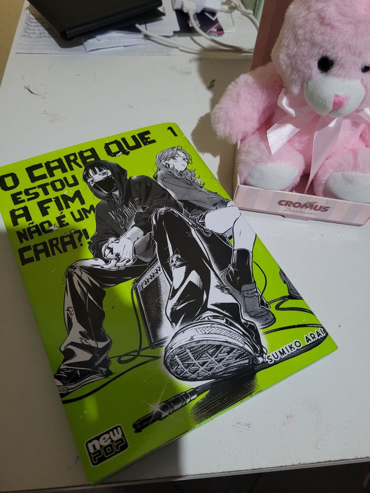 Na imagem há o mangá "O Cara Que Estou A Fim Não É Um Cara?!" volume 1, com capa vibrante em verde e ilustrações das personagens principais. Ao lado, um ursinho de pelúcia rosa sentado de lacinho no pescoço. Todos estão posto numa mesa branca.