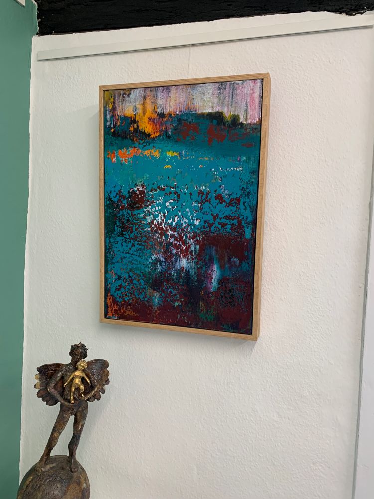 A multi-layered abstract painting on the wall in teal & dark magenta with touches of white, yellows and orange above and a sculpture below of an angel holding a child standing on a globe