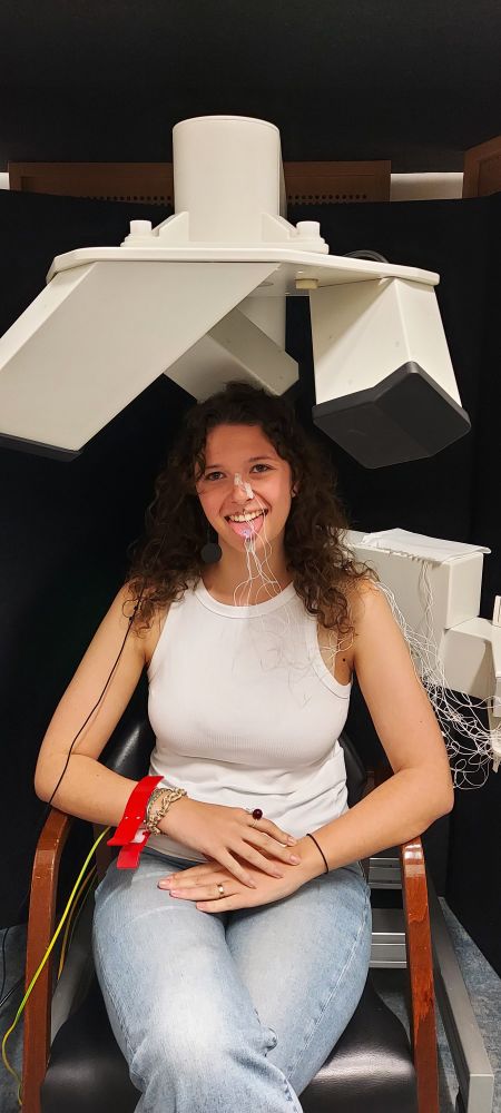 Une femme blanche brune en débardeur blanc et jean est assise sous une machine  EMA. Des capteurs sont collés sur son nez, ses lèvres et sa langue. Elle tire la langue et elle sourit.