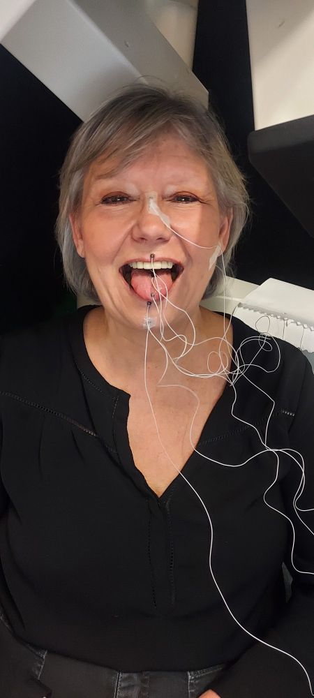 Une femme, la cinquantaine, de face, sourit en tirant la langue. Sur sa langue on distingue plusieurs bobines, atrachees à des fils qui partent vers la droite. D'autres bobines sont arrachées à son nez et ses lèvres. 