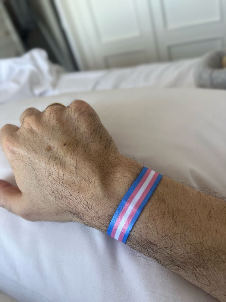 Se ve el antebrazo sobre una cama, el puño cerrado y en la muñeca una pulsera de tela con los colores del colectivo trans.
