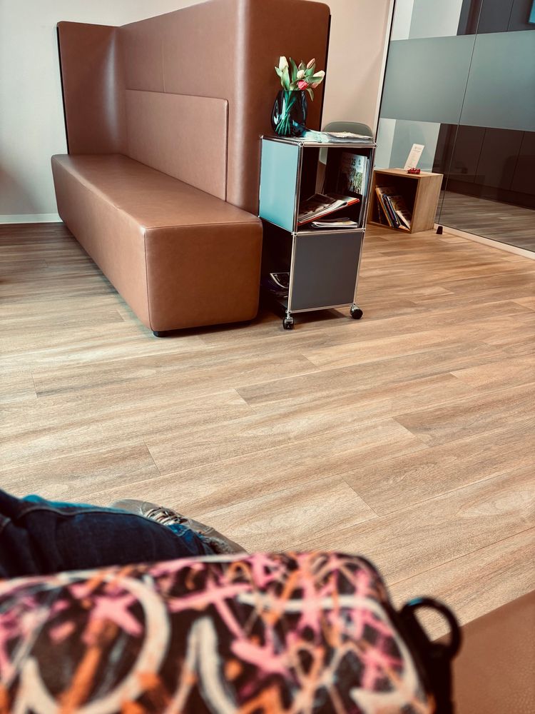 Ein Wartezimmer mit braunen Ledersitzmöglichkeiten, einem Beistelltisch mit einer Vase mit künstlichen Tulpen darin, Zeitschriften. Von mir ist nur ein kleiner Teil meiner Jeans, eines Stiefels und einer schwarzen Handtasche mit pink, orange, weißem Muster zu sehen.