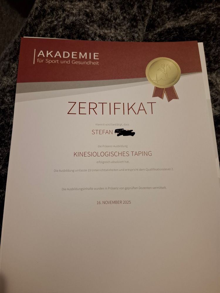 Das Bild zeigt ein offiziell aussehendes Zertifikat der "AKADEMIE für Sport und Gesundheit".
​Layout und Design
​Das Zertifikat liegt auf einer dunklen, möglicherweise grauen, flauschigen Oberfläche (wie ein Teppich oder eine Decke).
​Das Design ist professionell und verwendet die Farben Weiß, Dunkelrot/Bordeaux und Gold.
​Am oberen Rand befindet sich ein horizontaler Streifen in Dunkelrot und einem helleren Braunton.
​In der oberen linken Ecke steht in Großbuchstaben "AKADEMIE" und darunter in kleinerer Schrift "für Sport und Gesundheit".
​In der oberen rechten Ecke ist eine stilisierte goldene Medaille oder Plakette mit Bändern abgebildet, die als dekoratives Element dient.
​Inhalt des Zertifikats
​Die Hauptüberschrift, zentriert und in großer, serifenloser Schrift, lautet:
​ZERTIFIKAT
​Darunter folgt der Text, der die erfolgreiche Absolvierung einer Ausbildung bestätigt:
​"Hiermit wird bestätigt, dass"
​STEFAN [Name ist geschwärzt]
​"die Präsenz-Ausbildung"
​KINESIOLOGISCHES TAPING
​"erfolgreich absolviert hat."
​Details zur Ausbildung
​Zusätzliche Informationen zur Qualifikation werden genannt:
​"Die Ausbildung umfasste 19 Unterrichtseinheiten und entspricht dem Qualifikationslevel 2."
​"Die Ausbildungsinhalte wurden in Präsenz von geprüften Dozenten vermittelt."
​Datum
​Unten in der Mitte steht das Ausstellungsdatum: 16. NOVEMBER 2025.
