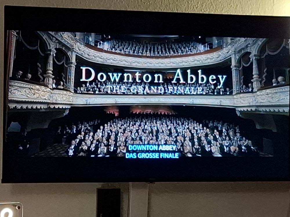 Das Bild zeigt eine Szene aus der britischen Fernsehserie Downton Abbey, wahrscheinlich von einem Fernseher oder Bildschirm abfotografiert.
Zentrale Szene: Zu sehen ist eine opulente Theaterszene, die das Innere eines großen Opernhauses oder Theaters zeigt, gefüllt mit einem festlich gekleideten Publikum. Die Kamera blickt von oben auf das Parkett und die Ränge.
Publikum: Das Publikum ist elegant gekleidet in Abendgarderobe (die meisten in Schwarz und Weiß), was der Zeit der Serie entspricht.
Schriftzug: In der Mitte des Bildschirms ist groß der Serientitel zu sehen, eingerahmt von dem Schriftzug:
Oben: "Downton Abbey"
Darunter, kleiner: "THE GRAND FINALE"
Im unteren Bereich, in der Mitte des Parketts, erscheint eine deutsche Übersetzung: "DOWNTON ABBEY DAS GROSSE FINALE"
Die gesamte Aufnahme wirkt feierlich und dramatisch, passend zu einem Serien- oder Film-Finale.