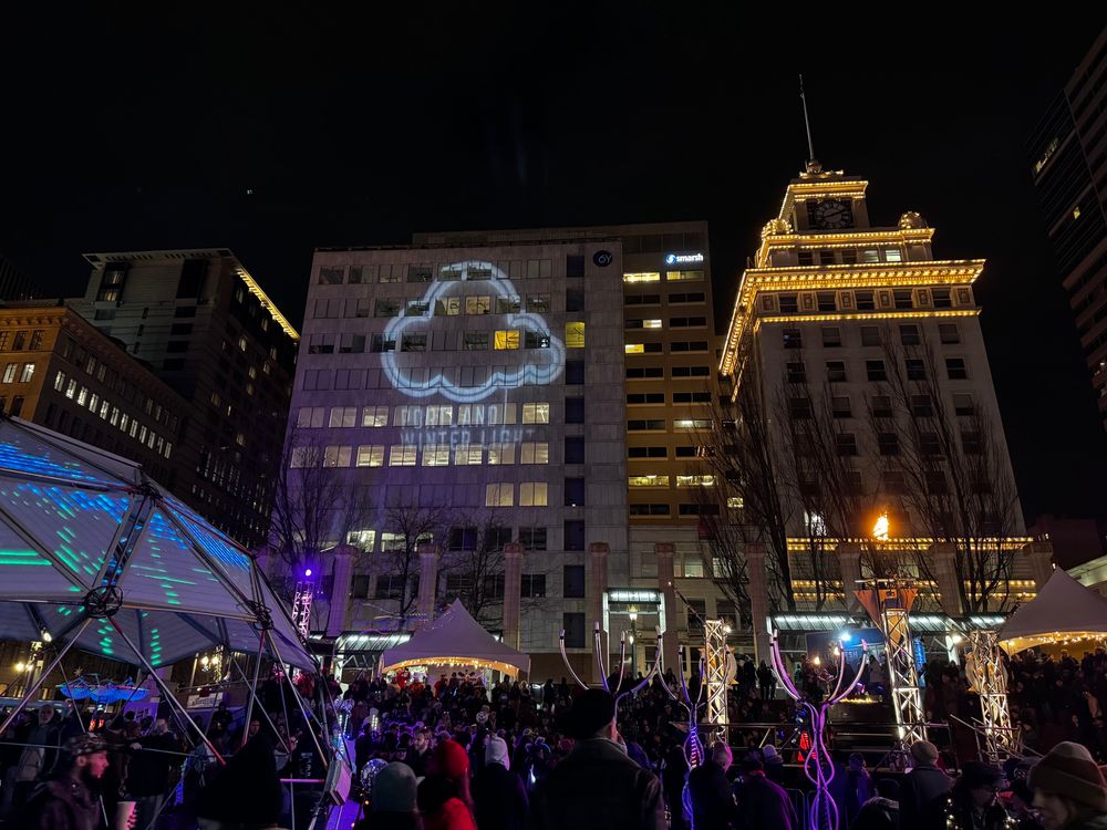 A large crowd at Pioneer Courthouse Square in Portland. A large geodesic dome made of LED panels is partially visible on the left while several smaller light sculptures occupy the Square. In the background the PDX Winter Lights logo is projected onto the side of an office building, while the facade of the high rise next to it is outlined with LED light strings.