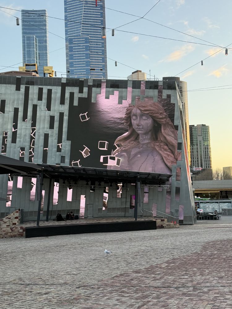 A giant marble face with pixel cubes flying towards her on a massive screen that has been split into shards across a building facade