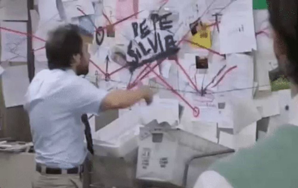 a man in a blue shirt and tie is pointing at a bulletin board with a spider on it .