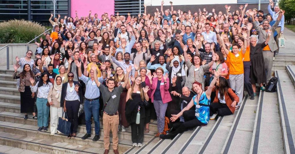 Speakers and delegates at the Critical Decade for Climate Conference on the UEA campus, hosted by the Tyndall Centre for Climate Change Research from 8-10 September 2025, stand smiling on the steps leading to the New Science Building, with their arms in the air