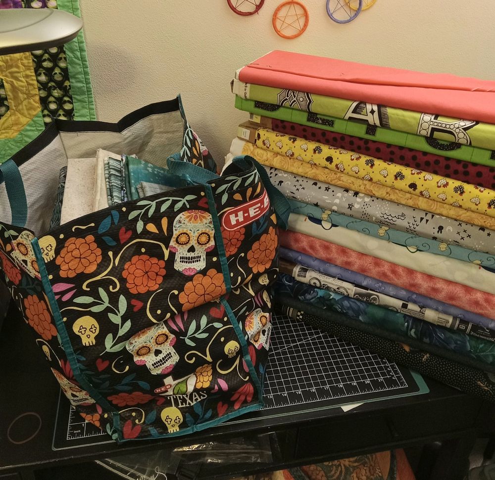 15 bolts of fabric yardage in a variety of colors and prints, next to a bag of folded fabric. The bag is about 14 inches high, 20 inches long, and very full.