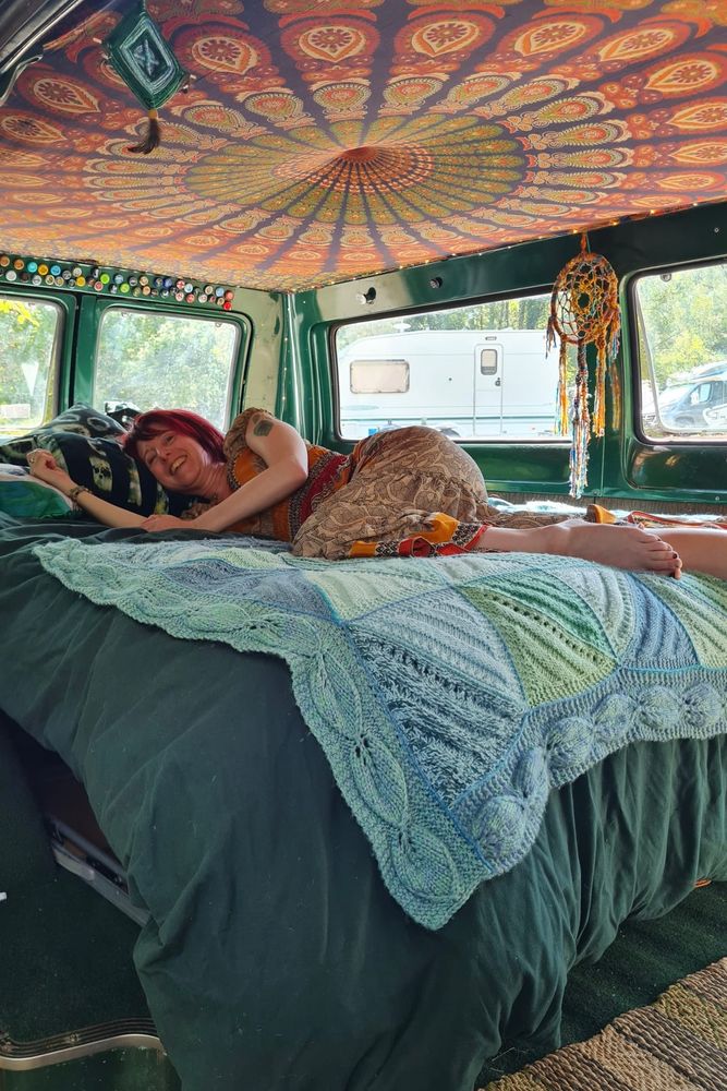 A white woman with short red hair and a green leaf tattoo on her arm is lying, smiling happily on a dark green bed with a knitted blue and green blanket. The bed is in a vintage campervan in a forest. It is decorated with a dream catcher, a woven eye of god, a collection of bottle caps and mandala throw covering the ceiling. The colours are all greens, blues and orange, giving a bohemian 70’s vibe. 