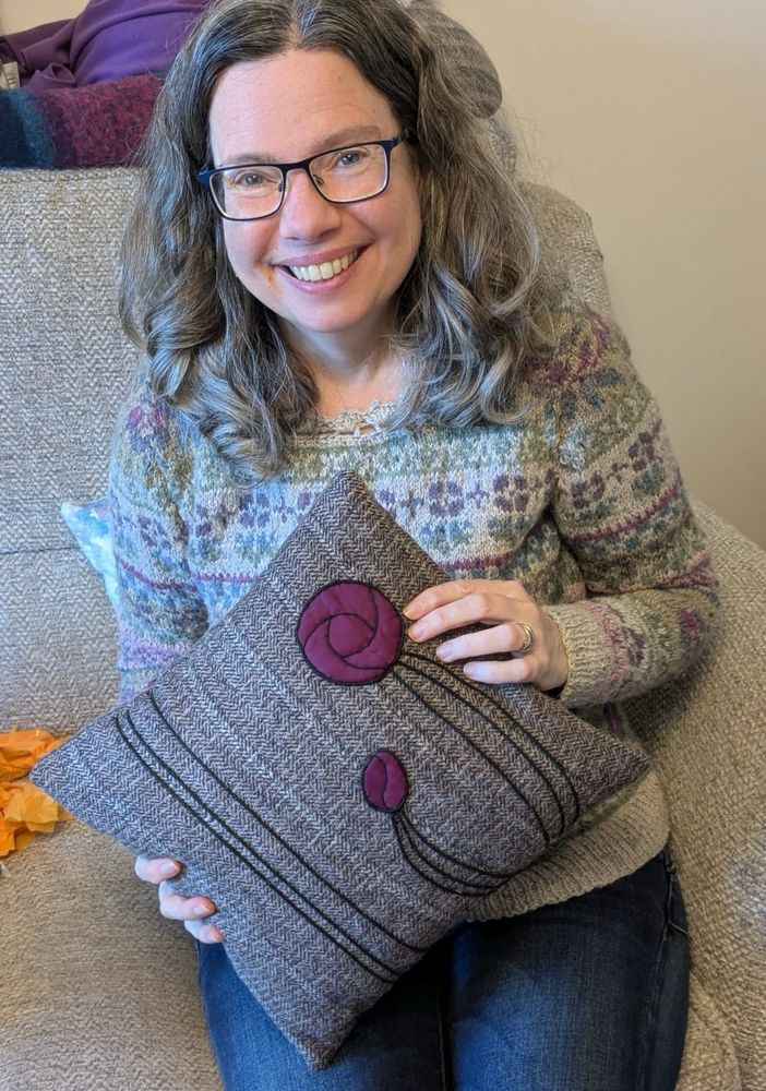 A white woman with a big smile and long curly hair is sitting on a sofa holding a cushion in tones of purple. The main fabric of the cushion is a heather coloured herringbone tweedy fabric. On this is an appliquéd design, in purple satin and black cord, inspired by Charles Rennie Macintosh’s Glasgow Rose stained glass piece. 