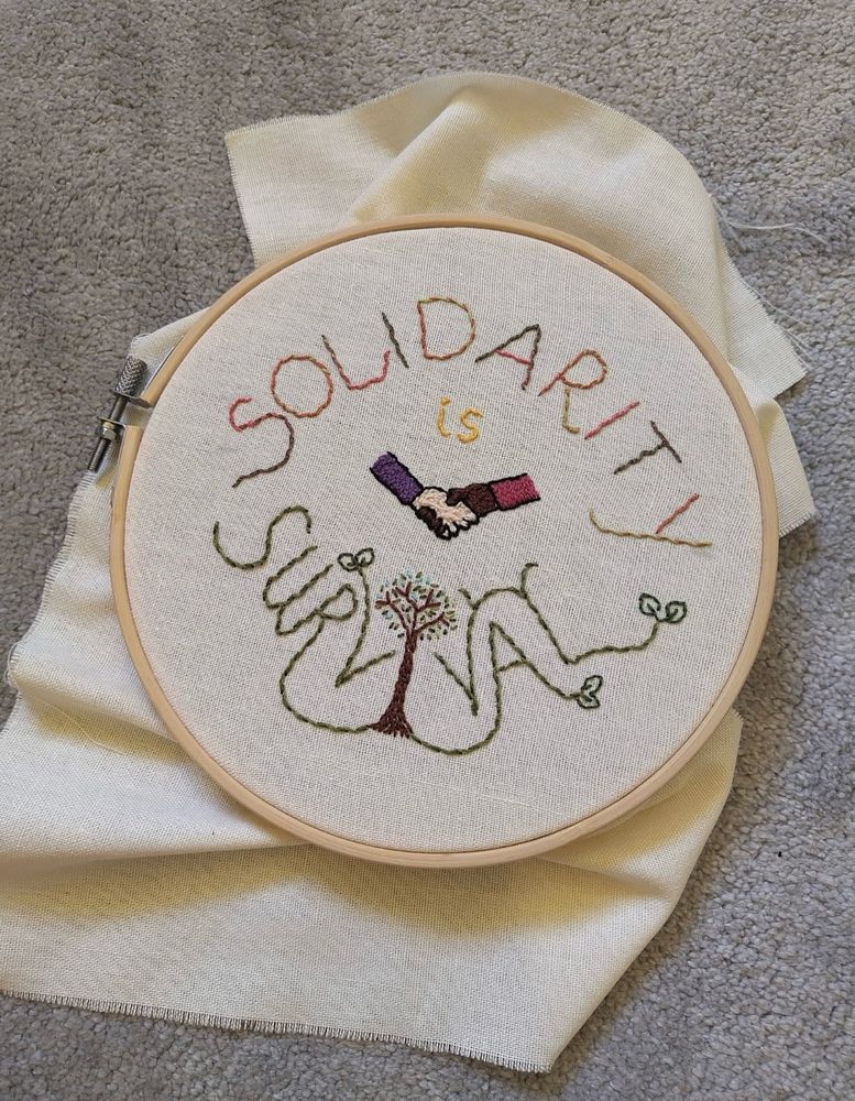 A hand sewn embroidery on cream fabric, still in its embroidery hoop. The neat sewing features the phrase ‘solidarity is survival’ forming a circular design. Inside the word survival is a tree whose roots become some of the letters. Other letters have tiny delicate leaves growing from them. At the centre of the design is an image of two arms holding hands, one black and one white, with each other. 