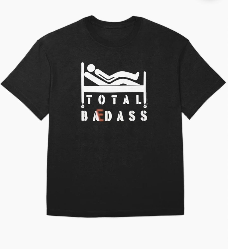 A black T-shirt with a white design by Paula J Knight depicting a person lying propped up in a hospital bed with the words Total Badass changed via a red letter E to Total Bedass. 