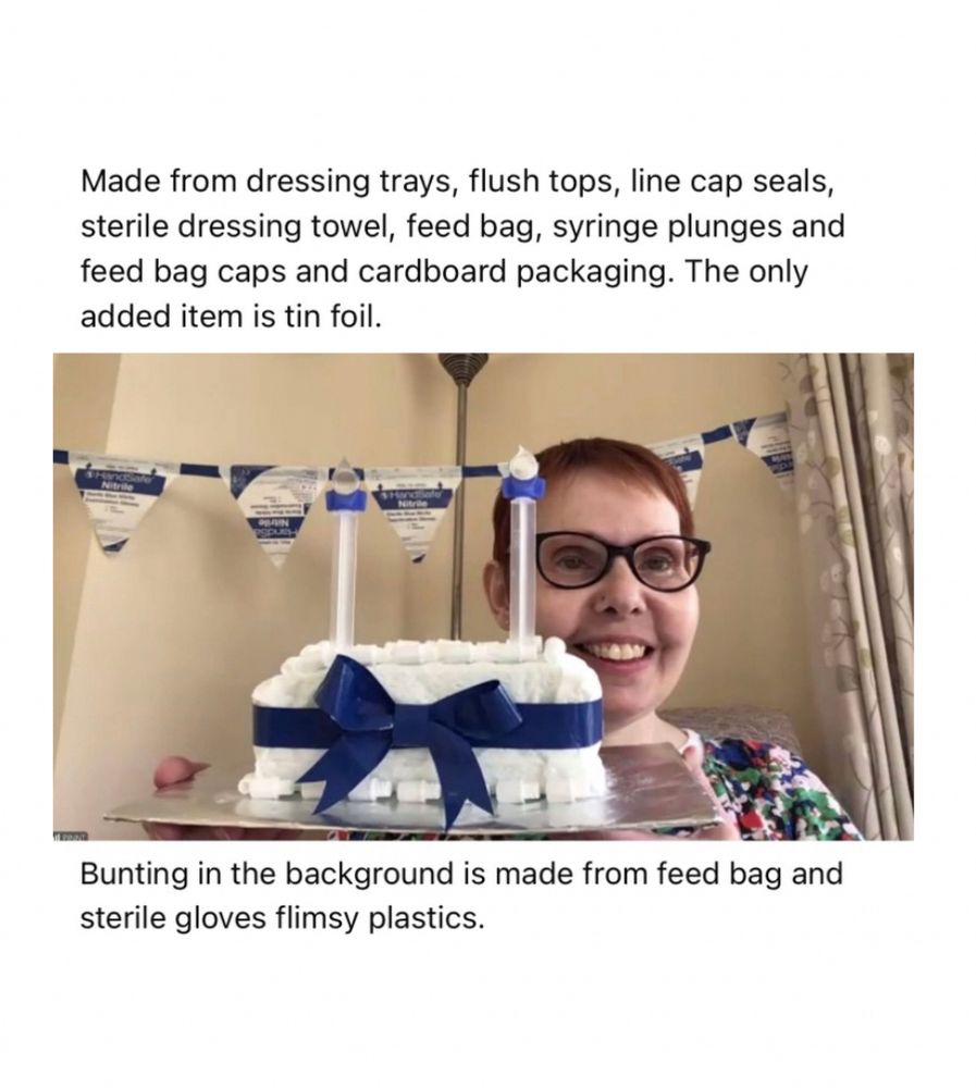 Sarah, a white woman with short dark hair, glasses and a big smile is holding a replica birthday cake with candles all created from blue and white dressing trays, flush tops, line cap seals, sterile dressing towel, feed bag, syringe plunges and feed bag caps, and cardboard packaging. The only added item is tin foil. Bunting in the background is made from feed bag and sterile gloves flimsy plastics.