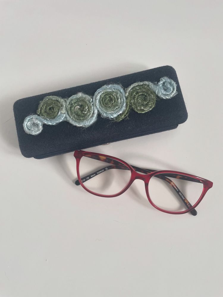 A pair of red-framed reading glasses with tortoise-shell patterned arms sits folded up next to a navy blue box-style rectangular glasses case. The top of the case is decorated with continuously joined spirals made by coiling yarn around and sticking it in place. The yarn changes colour, moving between a very pale blue to a sage green and is slightly shiny and very fibrous, being created from strands of recycled sari silk threads. 