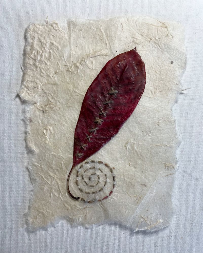 A red autumn leaf has been stitched to textured, torn-edged cream paper with cross stitches up the centre of the leaf using variegated brown sari silk thread. The same thread has been used to sew a running stitch spiral extending out from the leaf stem. 