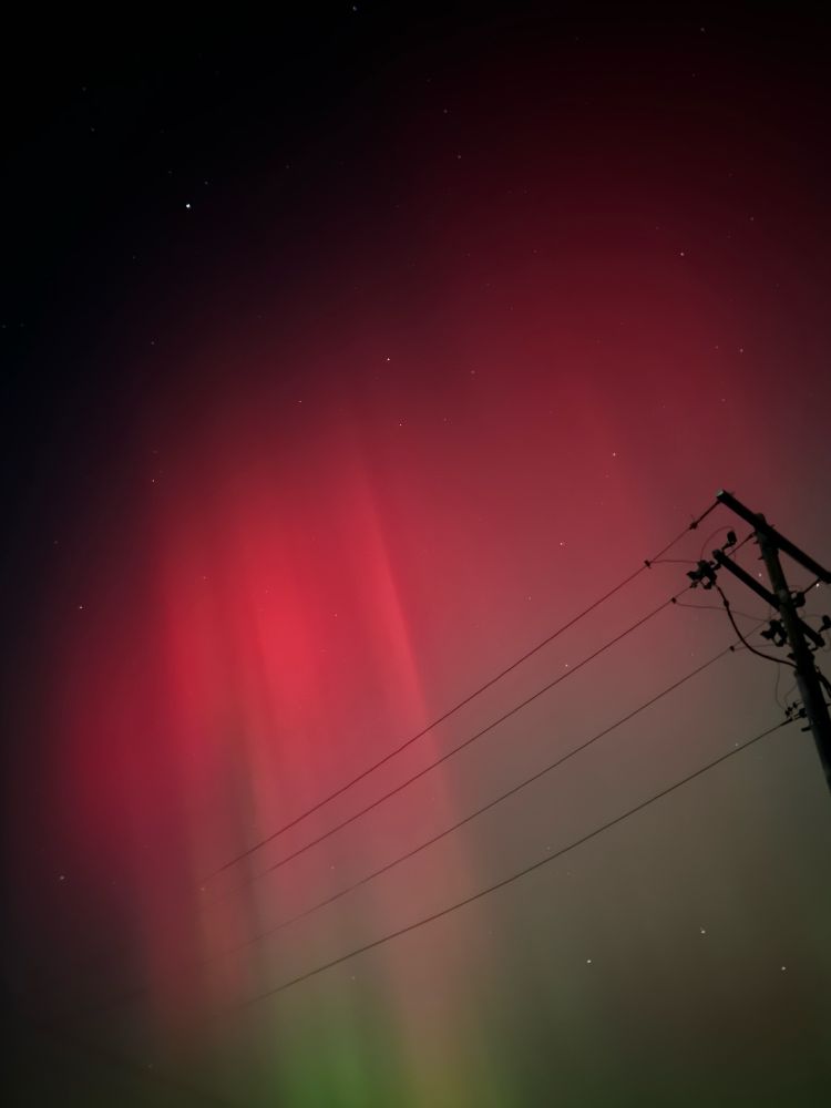 Aurora borealis in Illinois.  Vivid pink curtain of light fading in light green.  Electric lines dissect the picture as the power pole leans in to the right hand side of the photo.