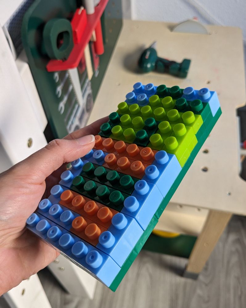 Ich halte eine flache, mit blauen, orangenen, dunkel und hellgrünen Bausteinen bebaute Platte in der Hand. Sie ist ca. 14x7cm groß. Im Hintergrund sieht man eine Spiel-Werkbank