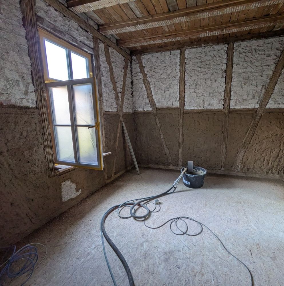 Ein Zimmer eines Fachwerkhauses im Rohbau. Man erkennt noch die Holzkonstruktion, aber die Gefache sind zum Teil bereits mit Lehm Unterputz bedeckt. Auf dem Boden liegen OSB Platten und darauf ein Eimer mit Putz. Links ist ein offenes Fenster zu sehen