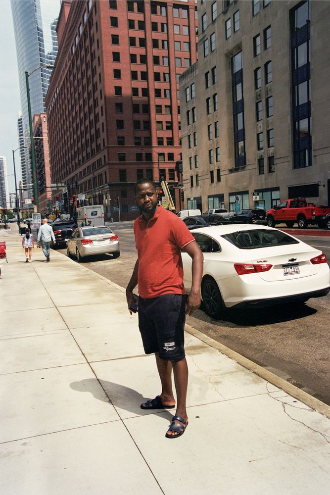 a middle-aged black man wearing a red polo and black shorts that go down to his knees. he's holding his cellphone and making a confident pose as he stares off to the right of the frame. the photo is in downtown chicago.
