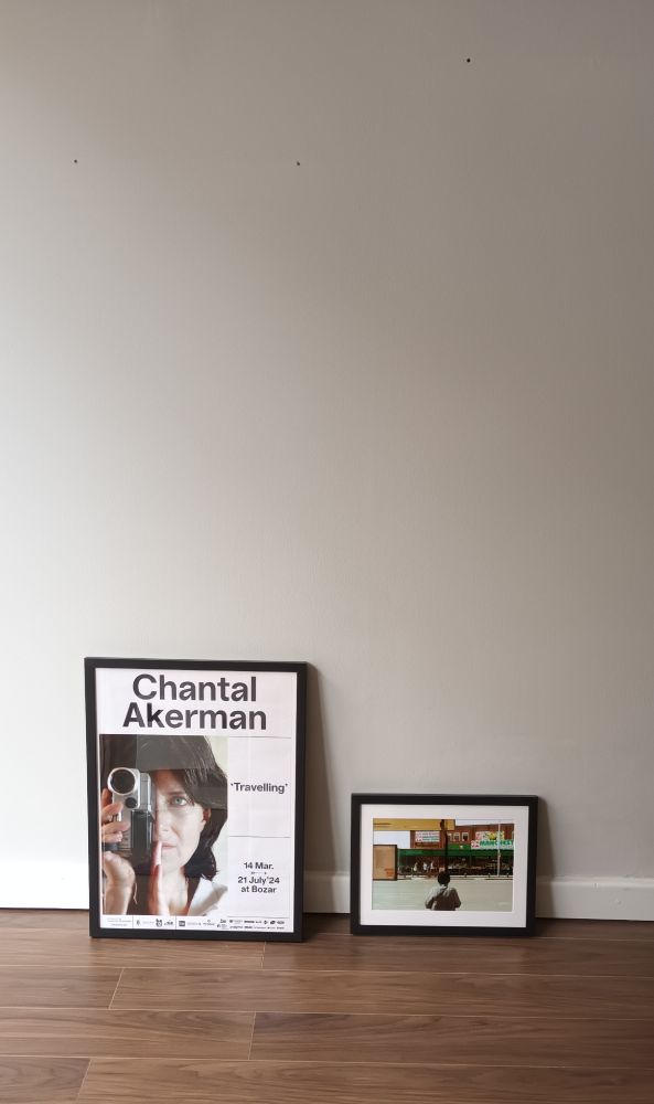 A framed picture of a Manchester grocery store and a framed photo of a Chantal Akerman poster, both placed on a wooden floor and against a wall.