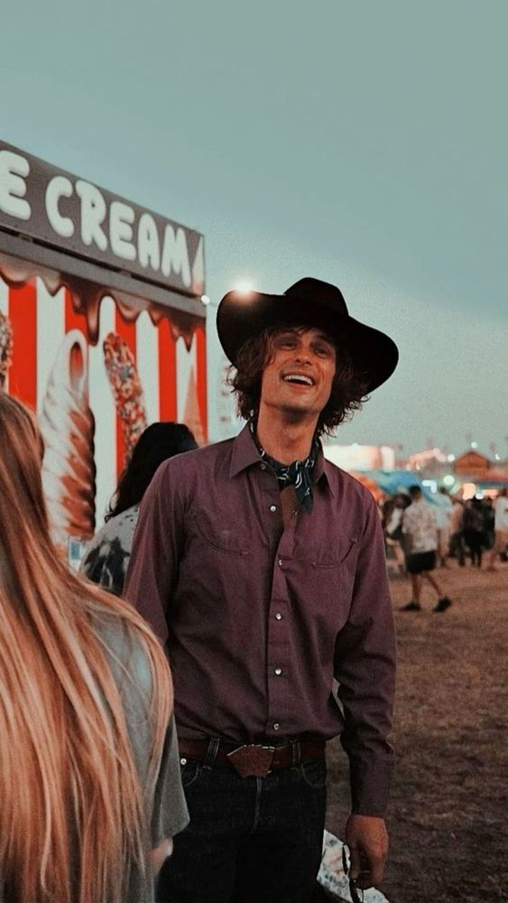 Matthew gray gubler in a purple shirt, cowboy hat, and bandana stands in front of an ice cream stand at a lively outdoor festival.