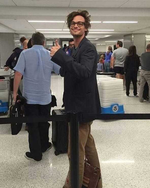 Matthew gray gubler giving a thumbs-up stands in an airport security line, surrounded by travelers and luggage.