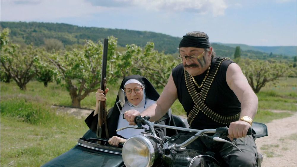 Frame from F.C. De Kampioenen: Kampioen Zijn Blijft Plezant (2013)
A rich industrialist is on the warpath dressed as Rambo and a nun is carrying his weapon.