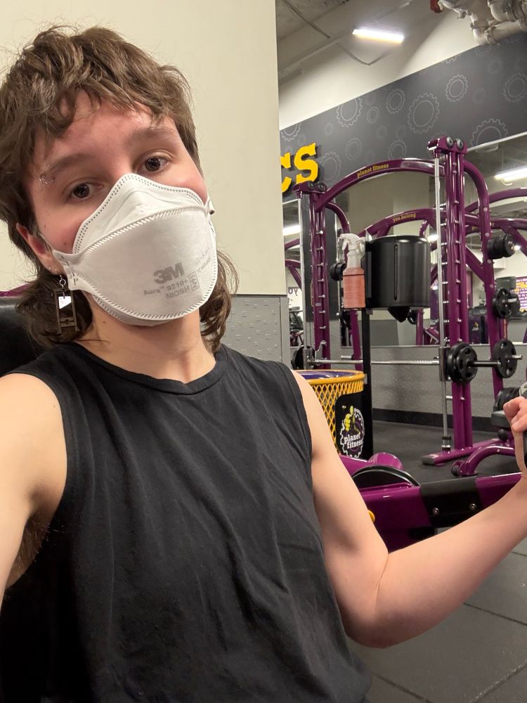 Sweetfern is a twenty-something lesbian with a mullet wearing an N95, a black tank, and guillotine earrings. They flex while sitting at a machine at planet fitness.