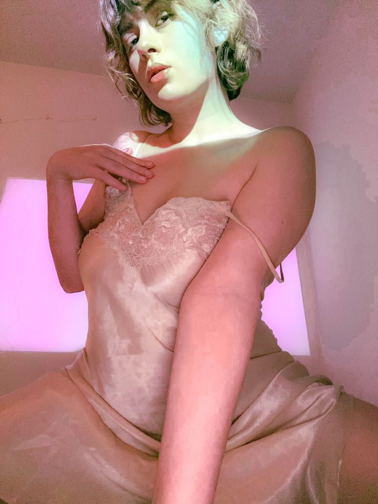 posed in front of the camera, gem's projector lights up her bust & background. gem's right hand is caressing her upper chest. left arm is posed in front of her, palm on her bed. her camisole nightgown strap is draped on her left shoulder