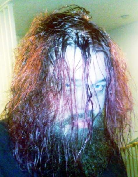 Lähikuva miehestä, jolla on pitkä, märkä ja punaruskea tukka sekä tuuhea parta. Hän katsoo intensiivisesti kameraan valaistuksessa, joka korostaa kasvojen kalpeutta.