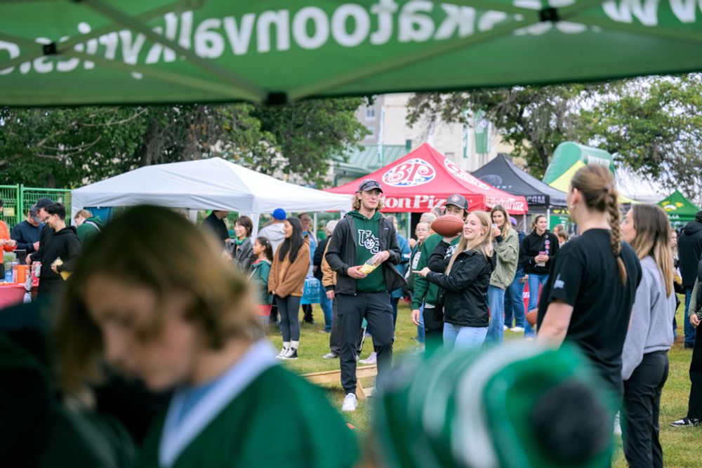 Group of enthusiastic University of Saskatchewan students cheering at an outdoor event, some holding banners and wearing school colors.

