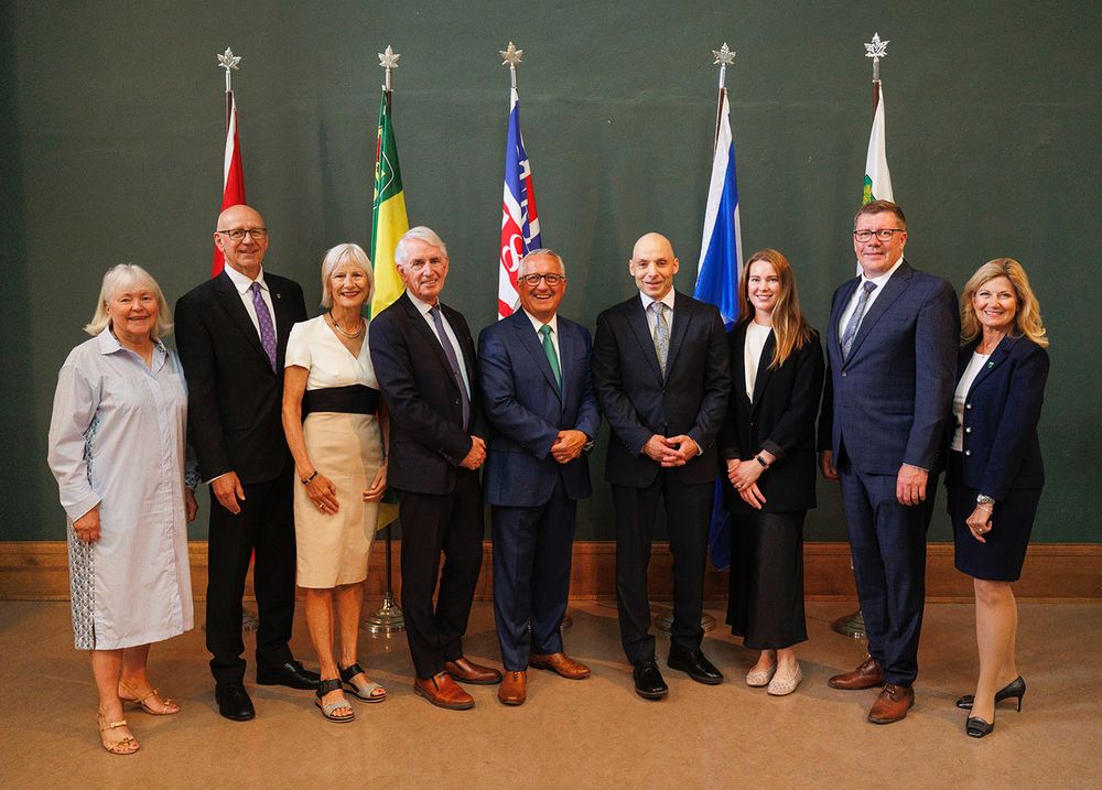 Group photo of eight dignitaries standing in front of their respective national flags, including the flags of Brazil, Canada, and others, in a formal setting.
