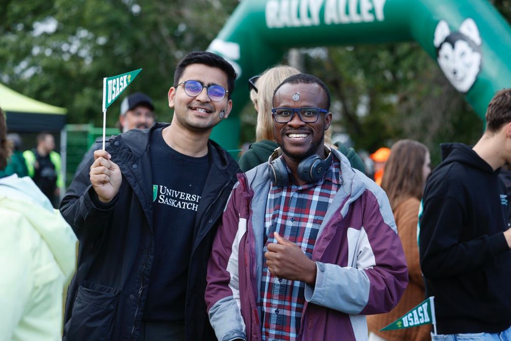 Two individuals smiling at an outdoor event, one holding a 'Yes!' flag, with a University of Saskatchewan sweatshirt visible. A rally setup with a green arch and a mascot in the background.
