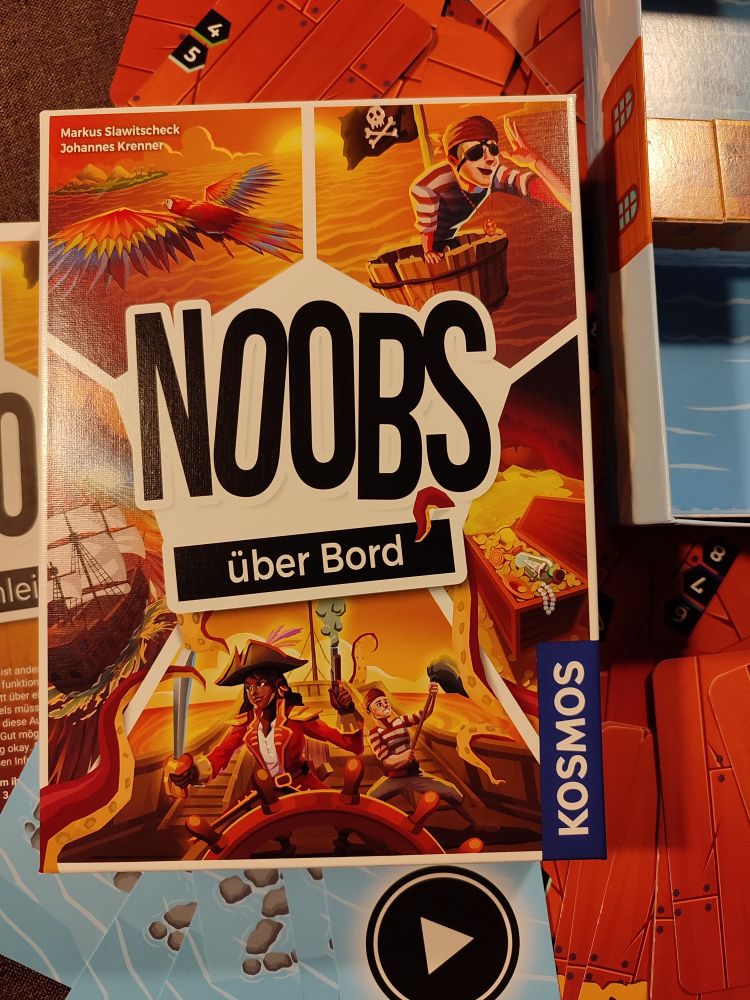 Spielschachtel von Noobs über Bord mit comichaften Piraten, Schatztruhe, Papagei und Segelschiff - Darunter Spielkarten