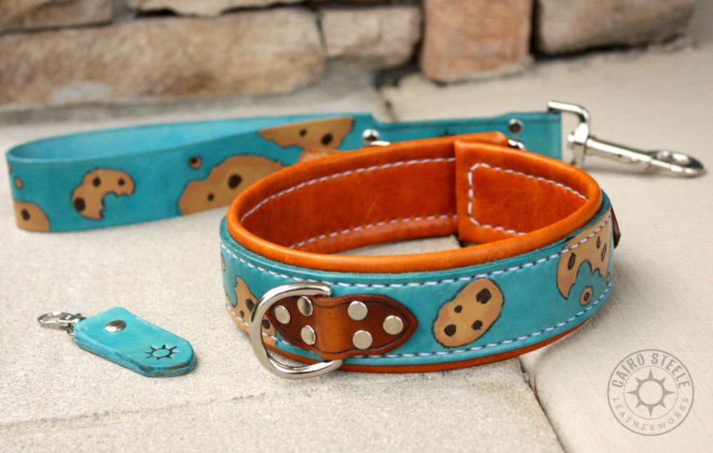 A collar and matching short leash. The collar has a light blue base strap with tooled and painted chocolate chip cookies, with a dark brown top strap, saddle tan liner, white stitching, and nickel hardware. The leash is light blue with cookies and a nickel clip.